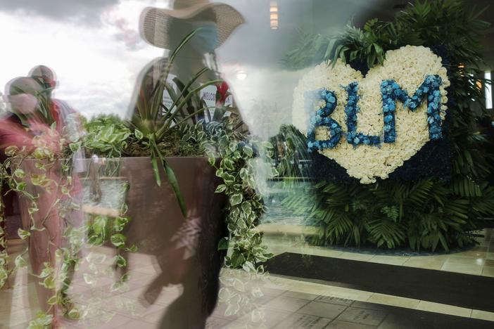 People are reflected in a window near a flower arrangement that includes the acronym for Black Lives Matter as they wait in line to attend the public viewing for George Floyd at the Fountain of Praise church on June 8, 2020 in Houston.