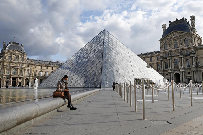 French President Emmanuel Macron is ordering nightly curfews for Paris and other large cities, hoping to control the coronavirus. A woman wears a protective face mask as she sits in the Louvre Museum's courtyard Wednesday in Paris.