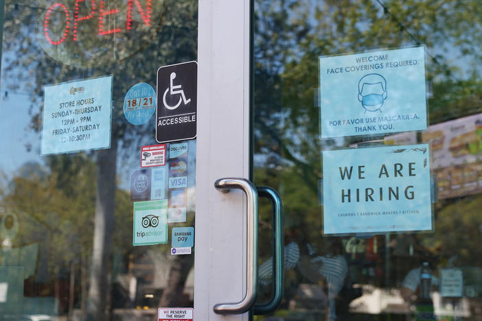 A "We are hiring" sign is paired with a pandemic-themed sign in the window of a store in Miami. Restaurants and other in-person businesses are looking to hire more workers at a time when some are wary of returning to work or are busy with caregiving.
