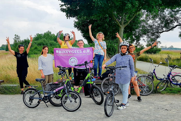 Volunteers and trainees with the group Bikeygees at a park in Berlin in July. The organization teaches refugee women in Germany how to ride bikes. Trainee Shapol Bakir-Rasoul, a refugee from Iraq, holds up a Bikeygees sign with founder Annette Krüger, right. Behind them in yellow is volunteer Shaha Khalef, a refugee from Iraq.