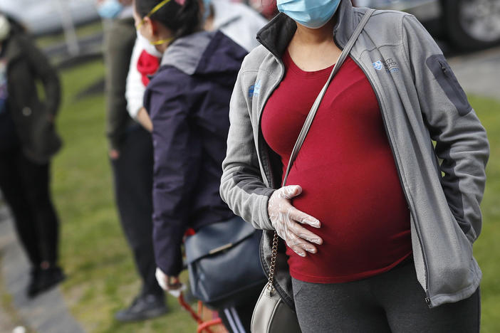 A pregnant woman wearing a face mask and gloves holds her belly as she waits in line for groceries at St. Mary's Church in Waltham, Mass., in May. The Centers for Disease Control and Prevention on Wednesday again urged all pregnant women to get the COVID-19 vaccine.