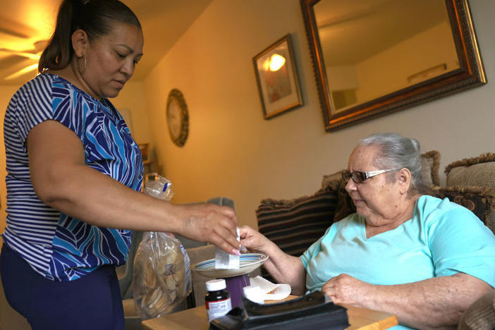 Expanded funds for in-home care can help seniors and disabled Americans stay in their homes. Here, Lidia Vilorio, a home health aide, gives her patient Martina Negron her medicine and crackers for her tea in May in Haverstraw, N.Y.