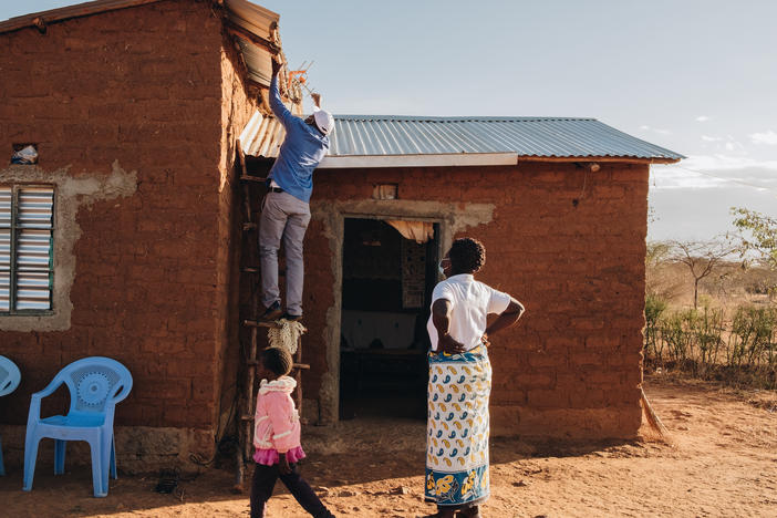 Winifred Muisyo, right, and her 5-year-old daughter, Patience Kativa, watch Stanlas Kisilu, left, as he installs a TV tuner on the roof of her home. The TV is connected to a solar panel provided by d.light, a company partially funded by climate financing from wealthier nations.