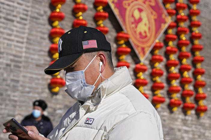 A man wearing a hat bearing an American flag, as well as a mask to help protect against the coronavirus, walks by a security guard near lanterns decorating remnants of a city wall in Beijing on Jan. 27.