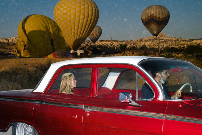 A woman and a man ride in a vintage car as hot air balloons float behind them in the region of Cappadocia, Turkey.