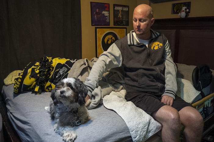 Jon Miller sits in his bedroom with his dog, Carlos, whom he received as a present for successfully completing cancer treatment a decade ago. Miller sustained severe brain damage, and requires the help of home health aides to continue living in his home.