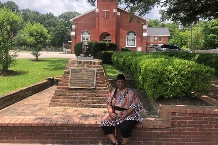 Vernetta Henson sits outside Union Baptist Church in Africatown. The church was started by Clotilda survivors in 1869. To her left is the bust of Cudjoe Lewis, one of the community's founder.