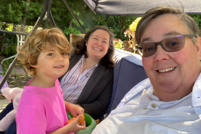 A selfie of Beth Kenny (foreground), their wife Adina (middle), and their child Vyla sitting in their backyard in Alameda, Calif. Since the lifting of COVID safety measures, Kenny and their family have had to pull back from indoor activities, and they struggle to explain why to Vyla.