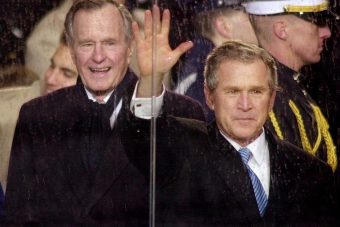 In this Jan. 20, 2001, file photo, standing in the rain, President George W. Bush waves as he watches his inaugural parade pass by the White House viewing stand in Washington, Saturday afternoon, Jan. 20, 2001. With him are his wife and first lady Laura Bush and his father, former President George H.W. Bush.