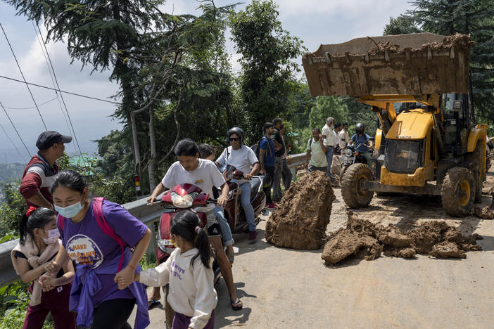 People rush past an earthmover clearing a road of a big rock that came down with mud and plant debris following intense monsoon rains in Dharmsala, Himachal Pradesh state, India, Sunday, Aug. 21, 2022.