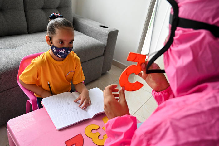 Giving a lesson at the home of a girl in Cali, Colombia, in August 2020, a teacher wears a biosecurity suit to prevent infection from the novel coronavirus. The "teacher at home" program aimed to help students stay in touch with teachers during pandemic school shutdowns.