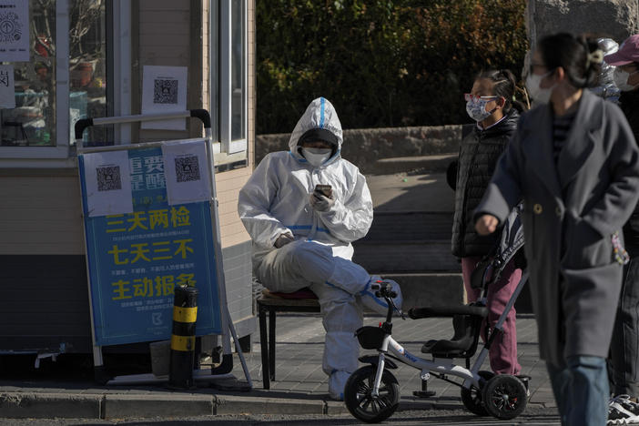 Residents walk by a security guard in protective suit browsing his phone at a main entrance gate to a neighborhood in Beijing, Thursday, Dec. 1, 2022.