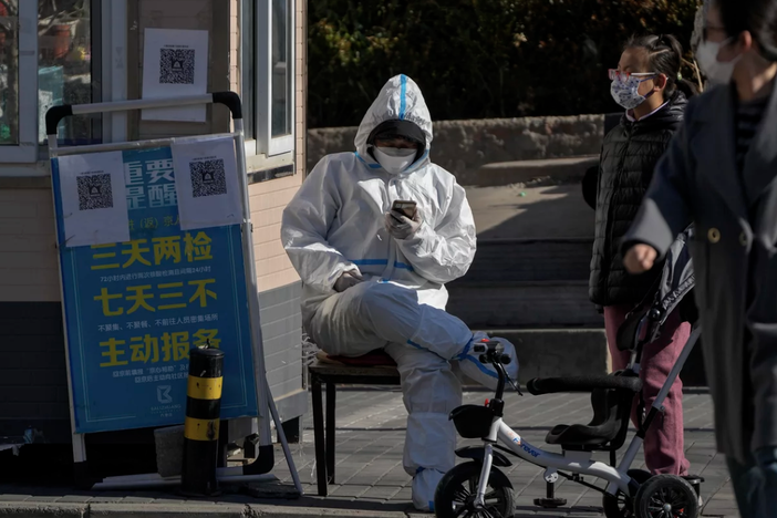 2022 年 12 月 1 日，星期四，北京某小区的主要入口，居民从一名身穿防护服的保安身旁走过，他正在浏览手机。