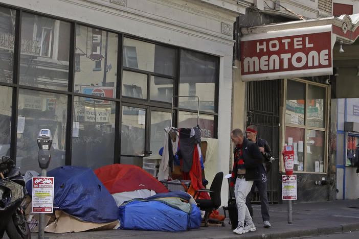 Pedestrians walk along the edge of a sidewalk to avoid tents and sleeping bags, on April 13, 2020, in the tenderloin area of San Francisco.