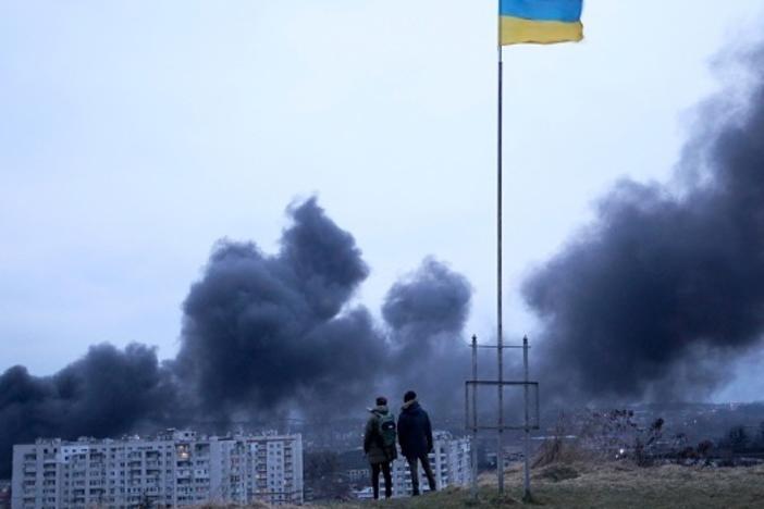 People standing near a Ukrainian national flag watch as dark smoke billows following an air strike in the western Ukrainian city of Lviv, on March 26, 2022.