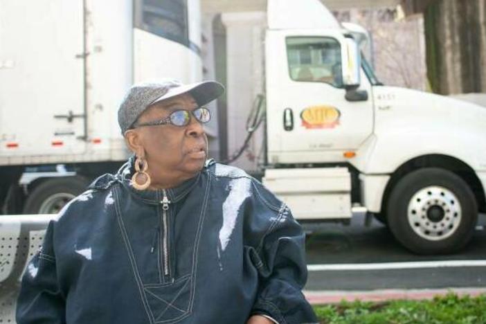 Community activist Margaret Gordon sits on a bench in West Oakland with the BART tracks behind her on March 4, 2022, as a semi-truck stops on 7th Street, on a popular trucking route to the nearby Port of Oakland.