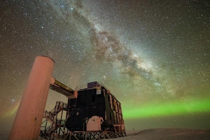 The IceCube Lab under a starry, night sky, with the Milky Way appearing over low auroras in the background.