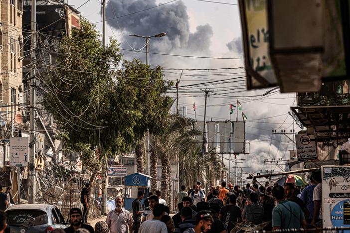 People walk along a street as a plume of smoke rises in the background during an Israeli strike on the Bureij refugee camp in the Gaza Strip on Thursday, as battles between Israel and the Palestinian Hamas movement continue.