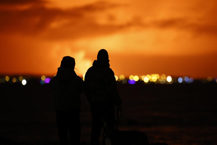 People watch as the night sky is illuminated caused by the eruption of a volcano on the Reykjanes Peninsula in southwestern Iceland seen from the capital city of Reykjavik on Monday, Dec. 18, 2023.