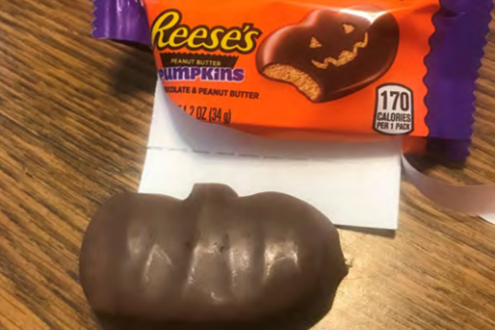 A Reese's Peanut Butter Pumpkin with no carved-out face next to the open wrapper with a jack-o'-lantern design.