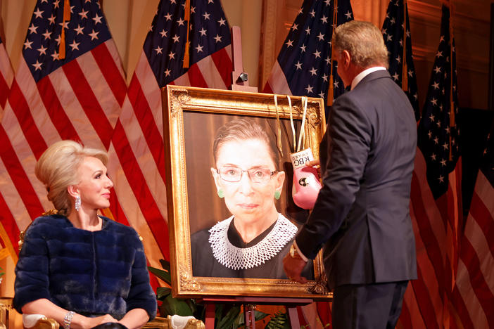 Sylvester Stallone hangs a pair of pink boxing gloves on a portrait of the late Supreme Court Justice Ruth Bader Ginsburg at the Justice Ruth Bader Ginsburg Woman of Leadership Award in 2022, as Julie Opperman, chair of the Dwight D. Opperman Foundation, looks on.