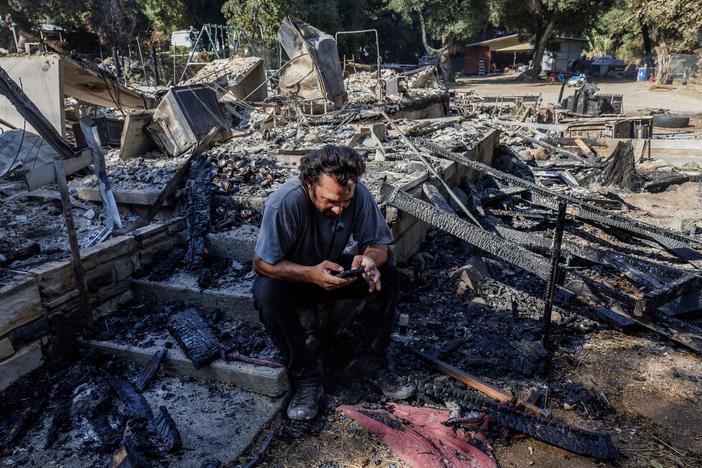 Garrett Keene sits on the front steps of his home in Lake Elsinore, Calif. that was destroyed in the Airport Fire, one of three major Southern California Fires so far this September. After evacuating with his family, Keene said he monitored the Watch Duty fire app to see where airdrops of fire retardant were happening as the fire approached, but none dropped on his neighborhood.