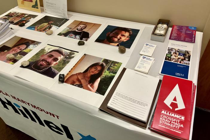 A table covered with a cloth featuring the Loyola Marymount Hillel logo in the front, and six portraits of hostages who were killed sitting on top of it.