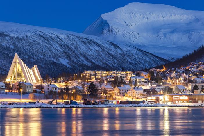 Dusk falls over snowy mountains that overlook the Arctic Cathedral and the houses mirrored in the icy fjord in Tromsø, Norway. Although this city is north of the Arctic Circle and experiences polar night for two months out of the year, its residents tend to see winter as a time of opportunity.