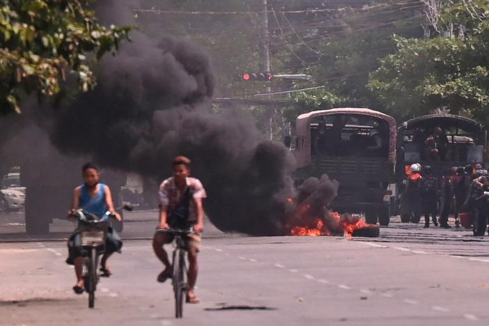 Cyclists pass police standing next to a burning makeshift tyre barricade, erected by protesters demonstrating against the military coup, in Yangon's South Okkalapa township on April 1, 2021.