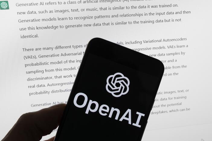 OpenAI's logo is on the screen of a mobile phone that's being held in front of a computer screen displaying output from ChatGPT.