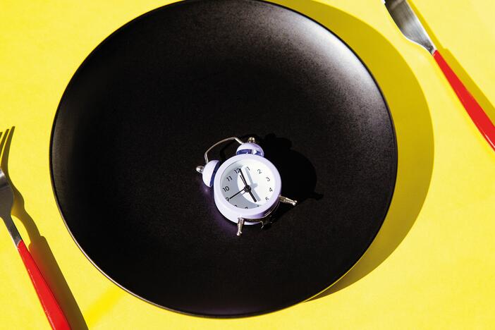 Small clock alarm on a black plate with red cutlery on yellow background.