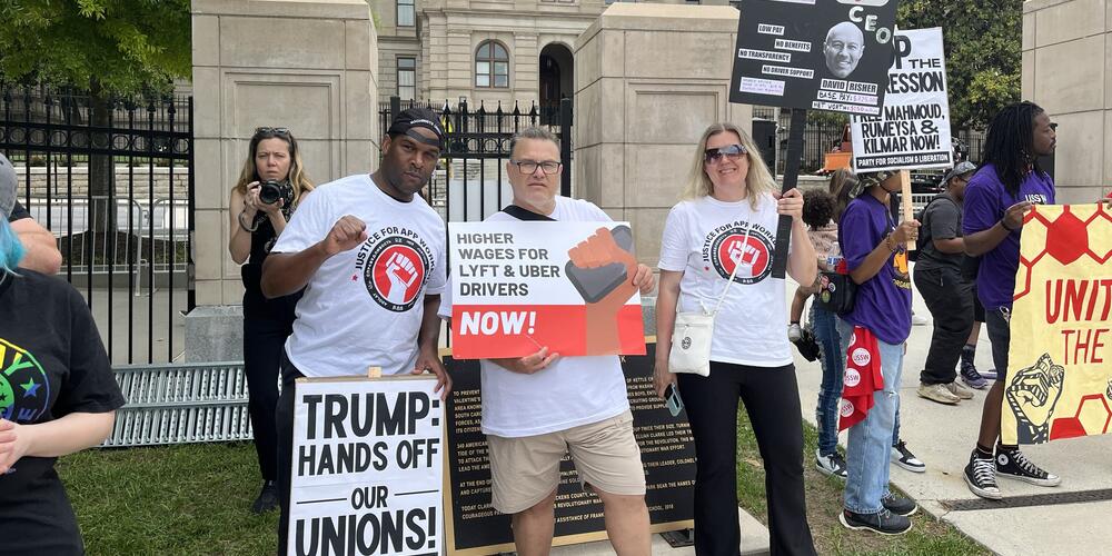 Three people in white shirts that say justice for app workers holding pro union signs.